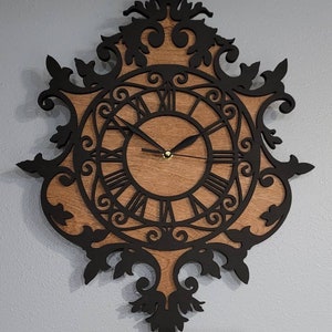 May include: A black and brown wooden wall clock with a decorative ornate frame and Roman numeral markings. The clock face is surrounded by a black filigree design.