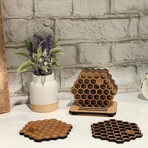 May include: Wooden honeycomb-shaped coasters and a decorative honeycomb structure. The coasters and structure are made of light brown wood. A small white vase with purple flowers is in the background.