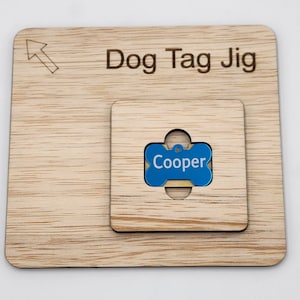 Op de afbeelding: Een houten hondenpenning jig met een uitsparing in de vorm van een bot. De jig is gelabeld "Dog Tag Jig" en heeft een pijl die omhoog wijst. Een blauwe hondenpenning met de naam "Cooper" is in de uitsparing geplaatst.