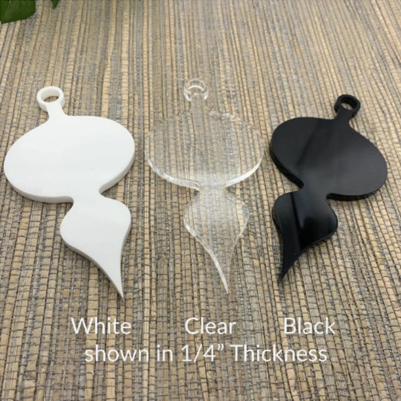 May include: Three acrylic ornaments in white, clear, and black. Each ornament has a rounded top with a hole for hanging and a stylized, elongated bottom. The text at the bottom reads "White, Clear, Black shown in 1/4" Thickness."