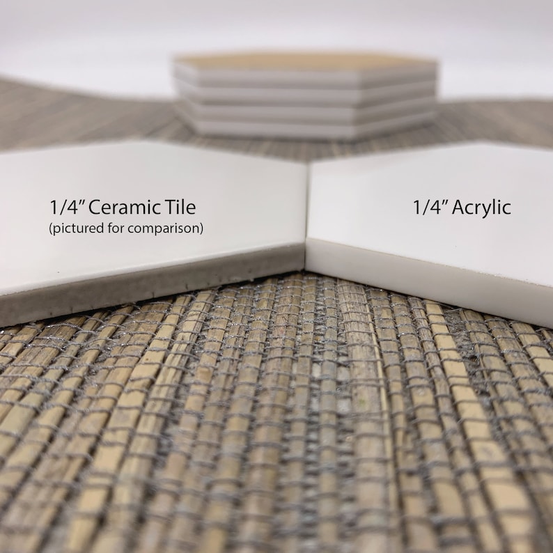 May include: Close-up comparison of two white hexagonal tiles. One is labeled "1/4" Ceramic Tile" and the other "1/4" Acrylic." The ceramic tile has a gray edge, while the acrylic tile has a clean white edge. A stack of similar tiles is in the background.