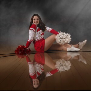 May include: A cheerleader in a red and white uniform, sitting on a wooden floor with a reflection. She holds white and red pom-poms. The uniform has the word "TEX" on the chest. She wears white sneakers and glasses.