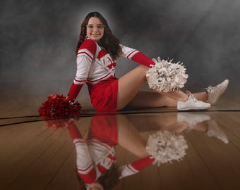 May include: A cheerleader in a red and white uniform, sitting on a wooden floor with a reflection. She holds white and red pom-poms. The uniform has the word "TEX" on the chest. She wears white trainers and glasses.