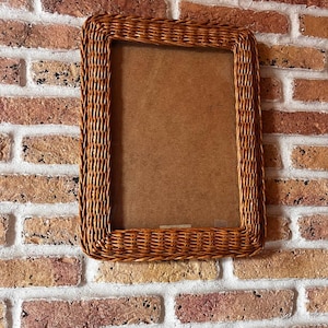 May include: A rectangular picture frame featuring a woven wicker border in a natural light brown finish. The frame has a smooth, flat inner edge and a textured, braided outer edge, surrounding a plain, solid brown backing material.