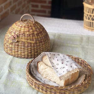 Könnte beinhalten: Ein geflochtener Brotkorb aus Weide mit passendem kuppelförmigen Deckel. Der Korb enthält zwei Brotscheiben, die in ein Stoff mit floralem Muster und Spitzenbesatz gewickelt sind. Korb und Deckel sind in Braun- und Gelbtönen gehalten.