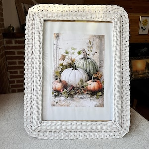 May include: White wicker picture frame with a rectangular shape. The framed artwork depicts a still life of pumpkins in shades of orange, white, and green, surrounded by autumn leaves. The frame has a woven texture.