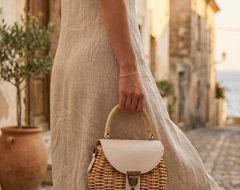 Può includere: Una borsa rotonda in vimini intrecciato, con patta e cinturino in pelle color crema. La borsa ha un manico in legno e una chiusura in metallo. È portata da una persona che indossa un abito di lino beige.