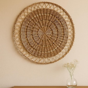 Puede incluir: Decoración mural redonda tejida en mimbre marrón natural. El diseño presenta círculos concéntricos y una superficie texturizada. El tejido está colgado en una pared neutra. Un pequeño jarrón de cristal con flores descansa sobre una superficie de madera.