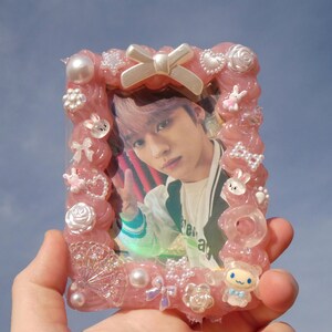 May include: A pink resin photo frame with decorative elements including bows, pearls, and small cartoon characters. The frame surrounds a photo of a person with pink hair. The frame is held against a blue sky background.