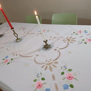 Puede incluir: Un mantel blanco con bordados florales en rosa, azul y verde. Tres velas encendidas en soportes ornamentados se colocan sobre la mesa, con una vela roja, blanca y blanca. La mesa está puesta para una ocasión especial.