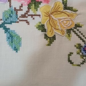 May include: Close-up of cream fabric with cross-stitch floral embroidery. The design includes a large yellow rose, blue leaves, and pink and purple flowers. The stitching is detailed and colourful.