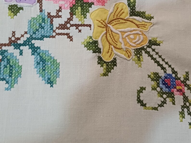 May include: Close-up of cream fabric with cross-stitch floral embroidery. The design includes a large yellow rose, blue leaves, and pink and purple flowers. The stitching is detailed and colourful.