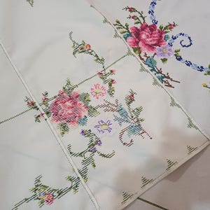 May include: Cream tablecloth with embroidered floral patterns. The embroidery includes pink, red, blue, and yellow flowers with green stems and leaves. The tablecloth has a stitched border.
