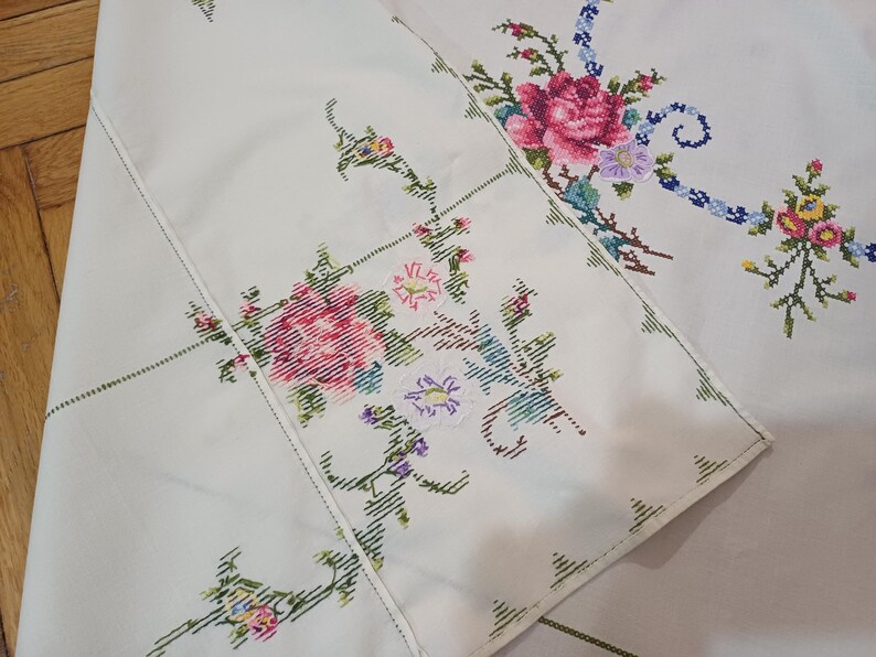 May include: Cream tablecloth with embroidered floral patterns. The embroidery includes pink, red, blue, and yellow flowers with green stems and leaves. The tablecloth has a stitched border.
