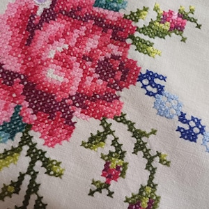 May include: Close-up of a cross-stitched floral design. The embroidery features a large pink and red rose with green leaves and stems. A blue and white chain-like pattern is also visible on the off-white fabric. The needlework is detailed and colourful.