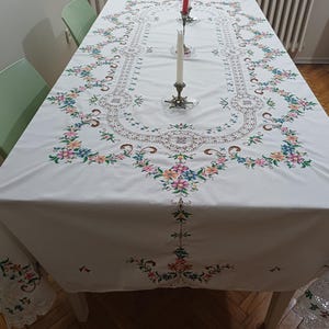 May include: A white rectangular tablecloth featuring intricate floral embroidery in shades of pink, blue, yellow, and green. The design includes a central openwork lace border and matching lace trim along the edges, with two tall candles in holders.
