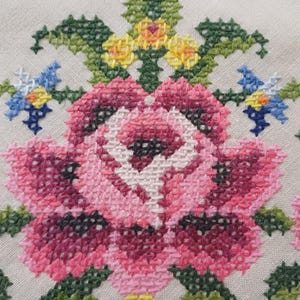 May include: Close-up of a cross-stitched floral design. The embroidery features a large pink rose with white and dark pink shading, surrounded by green leaves and smaller blue and yellow flowers. The background is a light beige fabric.