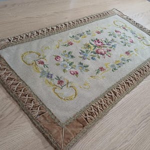 May include: Rectangular beige tapestry with a floral design in the center, featuring pink, blue, and yellow flowers. The tapestry has a brown velvet border with a decorative trim. The tapestry is placed on a light wood surface.