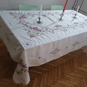 May include: White tablecloth with embroidered floral patterns in pink, red, and blue. The tablecloth is draped over a rectangular table. Three candle holders with lit candles are on the table.