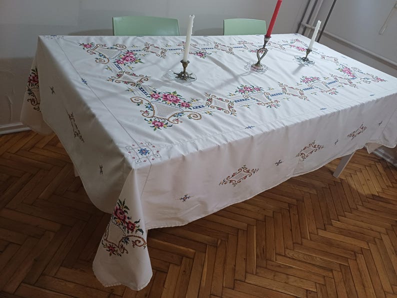 May include: White tablecloth with embroidered floral patterns in pink, red, and blue. The tablecloth is draped over a rectangular table. Three candle holders with lit candles are on the table.