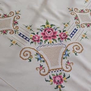 May include: Cream-colored tablecloth with intricate cross-stitch embroidery. The design features pink roses, blue and yellow flowers, and a basket motif. Decorative elements include geometric shapes and scrolling patterns.