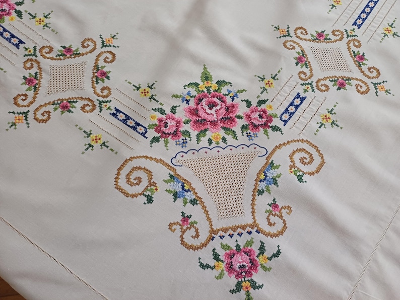 May include: Cream-colored tablecloth with intricate cross-stitch embroidery. The design features pink roses, blue and yellow flowers, and a basket motif. Decorative elements include geometric shapes and scrolling patterns.