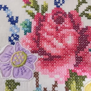 May include: Close-up of white fabric with cross-stitch embroidery. The design features a large pink and red rose, a purple flower, and blue and green floral accents. The embroidery is detailed and vibrant.