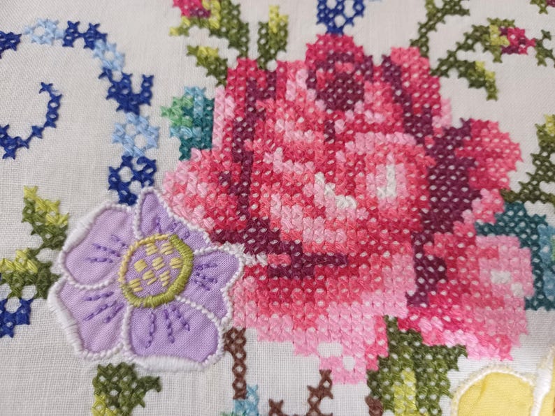 May include: Close-up of white fabric with cross-stitch embroidery. The design features a large pink and red rose, a purple flower, and blue and green floral accents. The embroidery is detailed and vibrant.