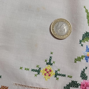 May include: Close-up of a beige fabric with colorful cross-stitch floral embroidery. The embroidery features pink, green, yellow, and blue flowers. A coin rests on the fabric, providing a size reference.
