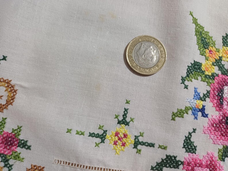 May include: Close-up of a beige fabric with colorful cross-stitch floral embroidery. The embroidery features pink, green, yellow, and blue flowers. A coin rests on the fabric, providing a size reference.
