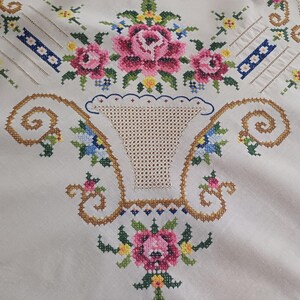 May include: Cream-colored tablecloth with intricate cross-stitch embroidery. The design features pink roses, blue and yellow flowers, and a basket motif. The embroidery is detailed with brown and green threads, creating a vintage aesthetic.
