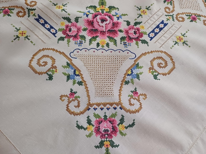 May include: Cream-colored tablecloth with intricate cross-stitch embroidery. The design features pink roses, blue and yellow flowers, and a basket motif. The embroidery is detailed with brown and green threads, creating a vintage aesthetic.