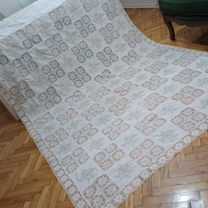 Könnte beinhalten: Eine weiße und beige Spitzentischdecke mit einem geometrischen Muster aus Quadraten und floralen Designs. Die Tischdecke ist über einen Tisch drapiert und zeigt ihre komplizierten Details und ihre zarte Textur. Die Ränder haben eine gewellte Bordüre.