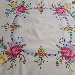 May include: Cream tablecloth with a cross-stitch floral design. The embroidery features pink roses, yellow and purple flowers, and green leaves. A blue ribbon-like border surrounds the floral arrangement.