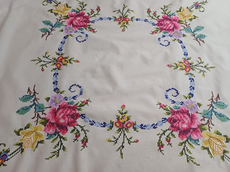 May include: Cream tablecloth with a cross-stitch floral design. The embroidery features pink roses, yellow and purple flowers, and green leaves. A blue ribbon-like border surrounds the floral arrangement.