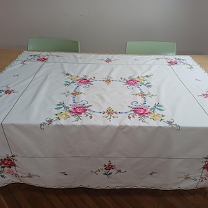 May include: A white tablecloth with colourful floral embroidery. The embroidery features pink, yellow, blue, and purple flowers arranged in a square pattern in the centre and along the edges. The tablecloth is rectangular and covers a wooden table.