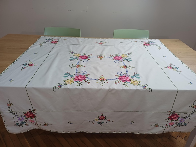 May include: A white tablecloth with colourful floral embroidery. The embroidery features pink, yellow, blue, and purple flowers arranged in a square pattern in the centre and along the edges. The tablecloth is rectangular and covers a wooden table.
