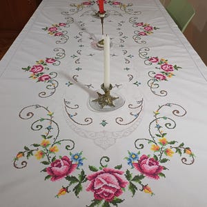 May include: White tablecloth with embroidered pink roses, green leaves, and blue and yellow flowers. A brass candlestick holds a white candle, and a red candle sits in a smaller holder. The table is set for a special occasion.