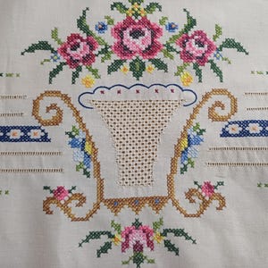 May include: Close-up of a cream-colored embroidered textile featuring a floral design. The design includes pink roses, blue and yellow flowers, and a basket-shaped motif. The embroidery is detailed with cross-stitch.