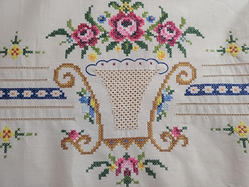 May include: Close-up of a cream-colored embroidered textile featuring a floral design. The design includes pink roses, blue and yellow flowers, and a basket-shaped motif. The embroidery is detailed with cross-stitch.