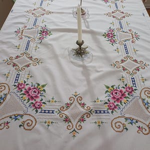 May include: White tablecloth with intricate floral embroidery in pink, green, blue, and gold. The design features repeating patterns of flowers, diamonds, and geometric shapes. A brass candle holder with a white candle sits on the table.