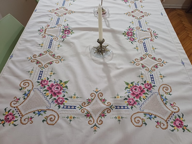 May include: White tablecloth with intricate floral embroidery in pink, green, blue, and gold. The design features repeating patterns of flowers, diamonds, and geometric shapes. A brass candle holder with a white candle sits on the table.