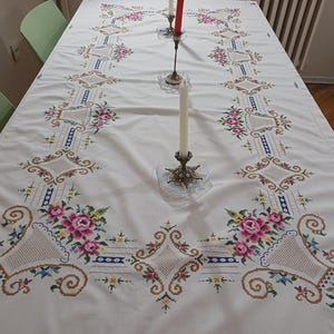 May include: A white tablecloth with intricate cross-stitch embroidery. The design features pink roses, blue accents, and geometric patterns. Two candle holders with lit candles are on the table, adding a touch of elegance to the dining setting.