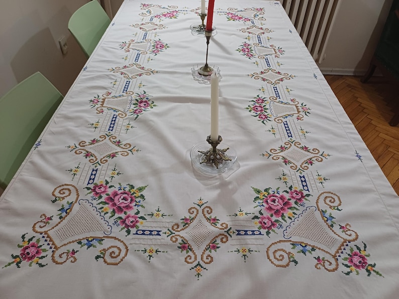 May include: A white tablecloth with intricate cross-stitch embroidery. The design features pink roses, blue accents, and geometric patterns. Two candle holders with lit candles are on the table, adding a touch of elegance to the dining setting.