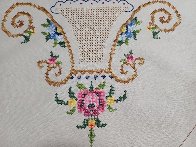 May include: Close-up of a cream-colored textile featuring cross-stitch embroidery. The design includes a central floral arrangement in pink, green, and yellow, with a basket-like shape above. The border is adorned with gold-colored swirls and blue and yellow flowers.