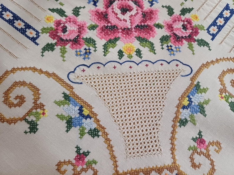 May include: Close-up of a cream-colored embroidered textile featuring a floral design. The embroidery includes pink roses, blue and yellow flowers, and a decorative border. The textile has a cut-out section with a lace-like pattern.