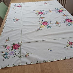 May include: A cream tablecloth with embroidered floral patterns. The embroidery includes pink, yellow, and purple flowers with green stems and leaves. The edges are trimmed with a green, scalloped border.