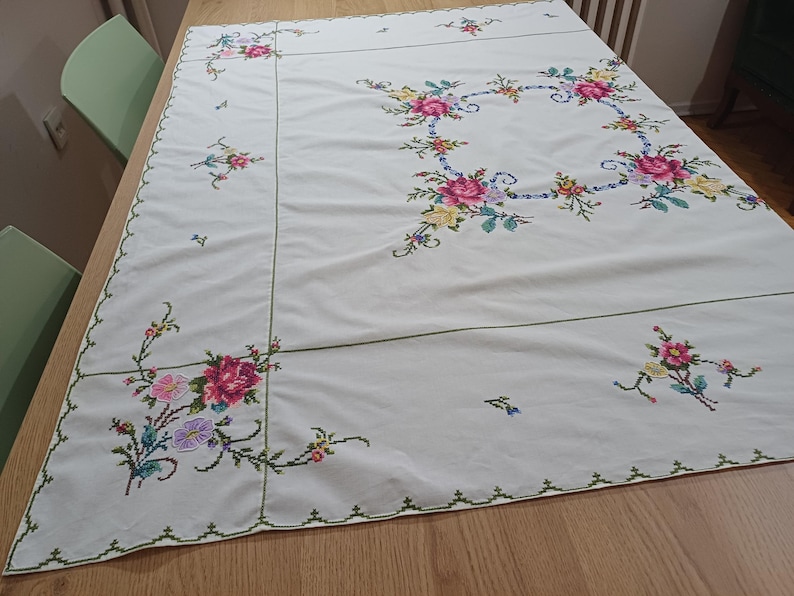 May include: A cream tablecloth with embroidered floral patterns. The embroidery includes pink, yellow, and purple flowers with green stems and leaves. The edges are trimmed with a green, scalloped border.