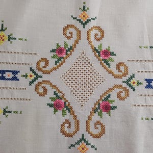 May include: Close-up of a cream-colored textile with intricate cross-stitch embroidery. The design features a central diamond shape with a lattice pattern, surrounded by brown swirls and pink and yellow floral accents. Blue and white bands are also present.