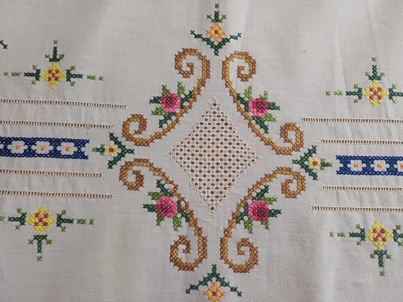 May include: Close-up of a cream-colored textile with intricate cross-stitch embroidery. The design features a central diamond shape with a lattice pattern, surrounded by brown swirls and pink and yellow floral accents. Blue and white bands are also present.